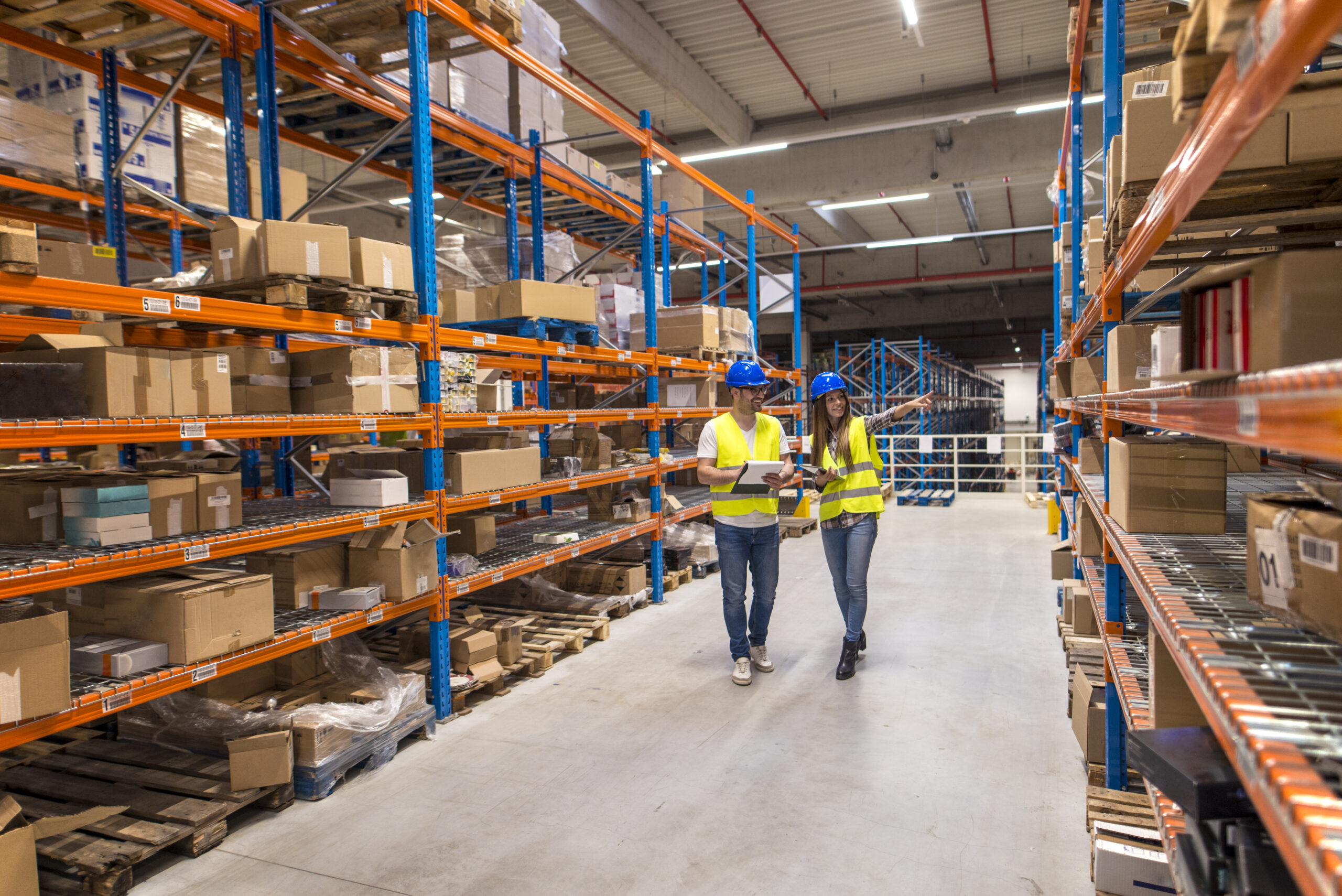 Two caucasian warehouse workers walking in distribution storage area discussing about logistics and organization.